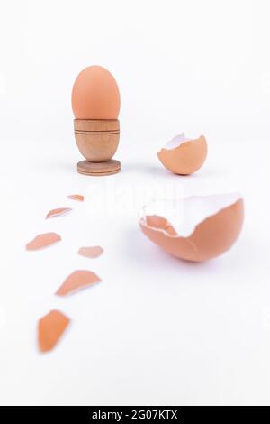 Tirer verticalement l'œuf de poule dans un ancien support d'œufs en bois et plusieurs coquilles d'œufs cassées sur une surface blanche. Minimalisme. Art conceptuel pour l'alimentation ou le produit animal Banque D'Images