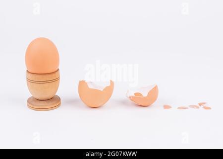 Œuf de poule horizontal dans un ancien support d'œufs en bois et plusieurs coquilles d'œufs cassées sur une surface blanche. Minimalisme. Art conceptuel pour l'alimentation ou produ animal Banque D'Images