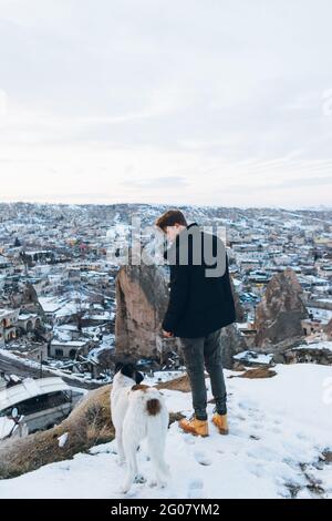 Vue arrière d'un jeune homme dans des vêtements chauds debout avec un chien fidèle sur une colline contre de petites maisons anciennes de grottes Dans la vallée enneigée au crépuscule en Turquie Banque D'Images