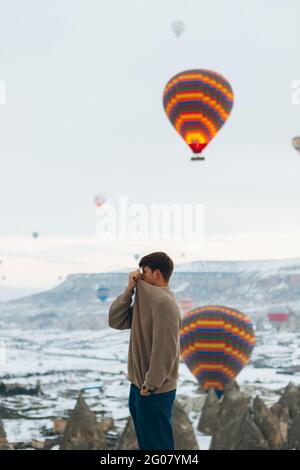 Vue latérale d'un homme qui regarde loin du paysage en se tenant debout contre les piliers de pierre insolites et les ballons d'air colorés en course survoler l'altitude brumeuse et enneigée Banque D'Images
