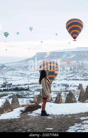 Vue latérale de la femme dans les vêtements chauds regardant pendant se tenant contre des piliers de pierre inhabituels et des ballons d'air colorés de course dans le ciel sur la neige brumeuse Banque D'Images