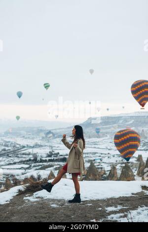 Vue latérale de la femme dans les vêtements chauds regardant pendant se tenant contre des piliers de pierre inhabituels et des ballons d'air colorés de course dans le ciel sur la neige brumeuse Banque D'Images