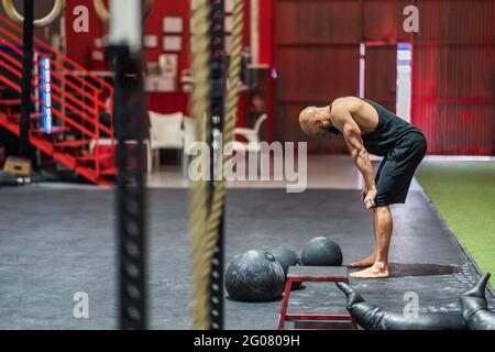 Vue latérale d'un chauve-homme musclé dans des vêtements de sport genoux et repos pendant l'entraînement fonctionnel dans la salle de sport spacieuse Banque D'Images