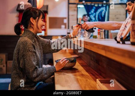 Vue latérale d'une femme asiatique satisfaite en chandail souriant tout en prenant la cuillère de l'ouvrier assis au comptoir en bois dans le bar ramen Banque D'Images