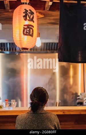 Vue arrière de la femme à cheveux noirs sans visage dans un chandail assis au comptoir dans un confortable bar ramen Banque D'Images