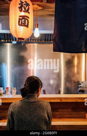 Vue arrière de la femme à cheveux noirs sans visage dans un chandail assis au comptoir dans un confortable bar ramen Banque D'Images
