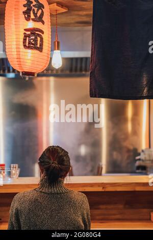 Vue arrière de la femme à cheveux noirs sans visage dans un chandail assis au comptoir dans un confortable bar ramen Banque D'Images