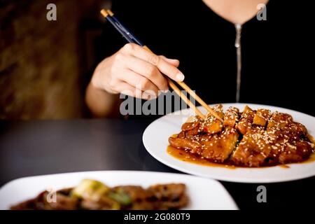 Crop femme dans une robe élégante assis dans le restaurant asiatique et manger du poulet savoureux et épicé préparé avec des baguettes Banque D'Images