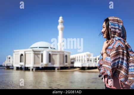 Vue latérale d'une jeune femme en foulard traditionnel, debout près de l'eau contre la célèbre Mosquée flottante ou la mosquée Al Rahma avec des murs en pierre blanche à Jedd Banque D'Images
