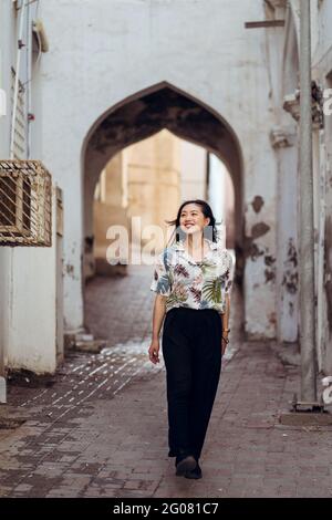 Tout le corps ravie jeune femme voyageur dans une tenue décontractée se promener le long de la rue étroite avec l'arcade du bâtiment ancien pendant la visite Souq. Muttrah Banque D'Images