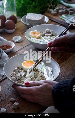 Personne non reconnaissable cultivée préparant des nouilles ramen fraîches cuites avec du tofu, des œufs et des légumes avec des baguettes sur une table en bois Banque D'Images