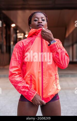 Jeune femme athlétique ethnique regardant l'appareil photo et la veste rouge de compression dans la rue Banque D'Images