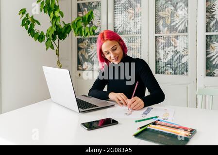 Jeune créatrice pensive femme designer de mode dans une tenue décontractée et lunettes assises à la table avec des gadgets et dessin avec crayons colorés Banque D'Images