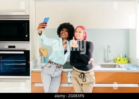 Des amies multiraciales stylées et joyeuses dans une tenue décontractée cuisine et prise de selfie avec téléphone portable Banque D'Images