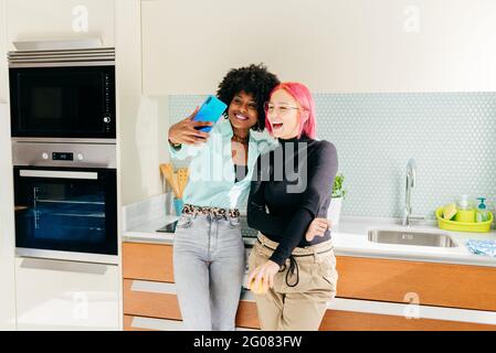 Des amies multiraciales stylées et joyeuses dans une tenue décontractée cuisine et prise de selfie avec téléphone portable Banque D'Images