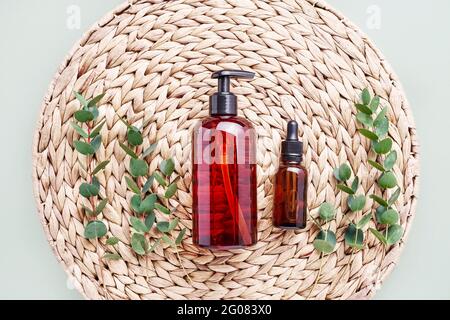 Bouteille compte-gouttes d'huile essentielle ou de sérum, branches d'eucalyptus et bouteille de distributeur sombre de gel douche sur fond en rotin. Concept beauté et SPA. Banque D'Images