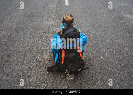 Vue arrière de la femme tranquille en blouson bleu vif avec sac à dos assis sur la route et regardant loin Banque D'Images