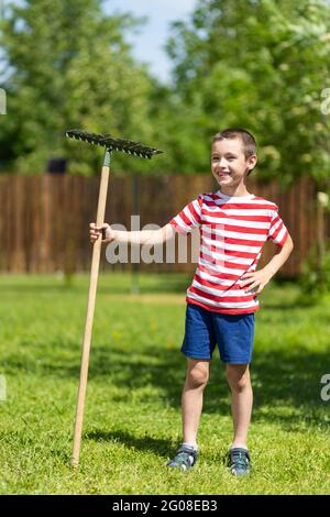 Un petit garçon joyeux tient un rake dans sa main, prêt à travailler dans le jardin d'une maison de campagne. Banque D'Images