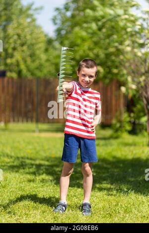 Un petit garçon joyeux tient un rake dans sa main, prêt à travailler dans le jardin d'une maison de campagne. Banque D'Images