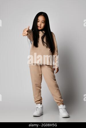 Positif asiatique fille dans vert décontracté vêtements de sport pantalon en coton, sweat à capuche, jersey et baskets se tient gesturant le pouce vers le haut Banque D'Images