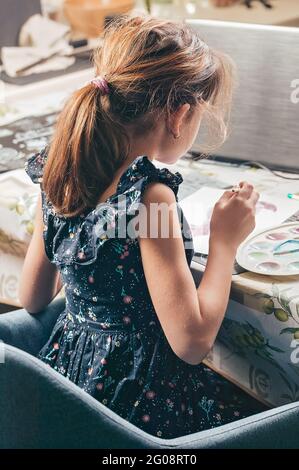 Une fille s'assoit et peint à la table. Elle regarde une leçon en ligne. Cours et études pendant la pandémie COVID-19. Vue arrière. Idées pour activiti Banque D'Images