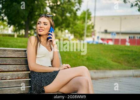 Bonne fille qui appelle sur un téléphone portable regardant loin assis sur un banc dans la rue. Jeune fille blonde ayant une conversation détendue dans un parc. Photo de haute qualité Banque D'Images