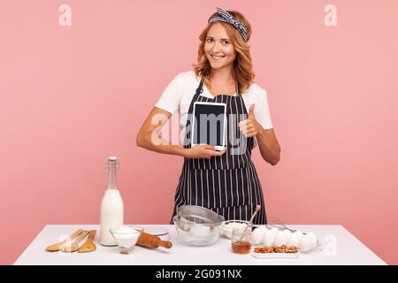 Portrait d'une jeune femme adulte attrayante dans une boulangerie à tablier, tenant une tablette vierge à la main et montrant le pouce vers le haut, regardant l'appareil photo avec le sourire. Po Banque D'Images