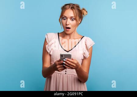 Belle jeune femme adulte étonnante en robe d'été, en utilisant un smartphone et regarder la vidéo choquante, la lecture de message avec l'expression stupéfiante. Intérieur s Banque D'Images