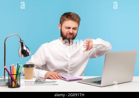 Je n'aime pas ! Homme barbu travailleur de bureau assis sur le lieu de travail montrant le pouce vers l'écran d'ordinateur portable, exprimant sa désapprobation, insatisfait de l'étalonnage vidéo Banque D'Images