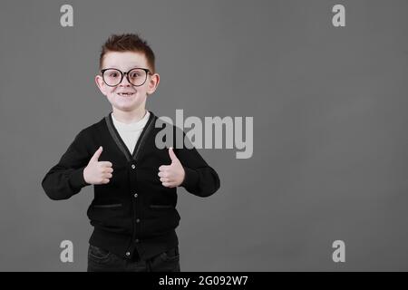 Comme. Portrait d'un jeune écolier heureux avec des lunettes souriant à l'appareil photo et faisant un geste de pouce vers le haut, montrant un signe d'approbation sympa d'accord. Studio d'intérieur isolé sur fond gris. Banque D'Images