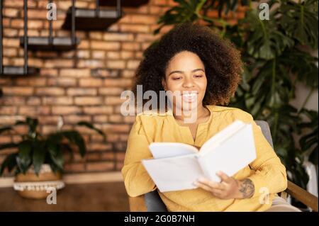 La jeune femme afro-américaine qui se repose sur un livre assis sur un fauteuil dans un salon confortable, apprécie le nouveau BEST-seller, passe un week-end à la maison Banque D'Images
