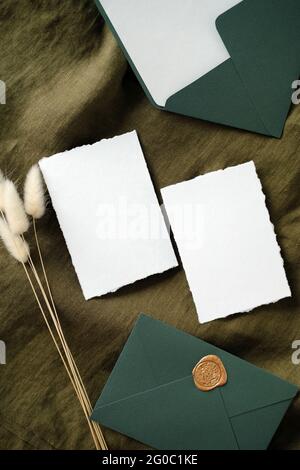 Cartes d'invitation de mariage élégantes et enveloppes sur tissu vert avec fleurs séchées. Pose de la feuille, vue de dessus, espace de copie. Banque D'Images