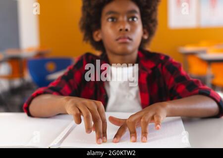 Un écolier américain africain aveugle assis à un bureau dans une salle de classe lisant un livre en braille avec les doigts Banque D'Images