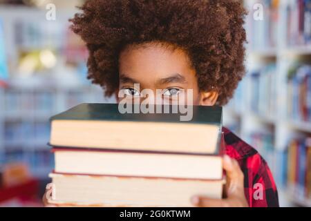 Portrait d'un écolier afro-américain portant une pile de livres dans la bibliothèque scolaire Banque D'Images