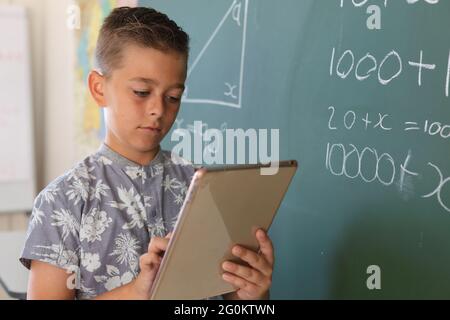 Garçon caucasien debout au tableau de surveillance en classe et utilisant une tablette pendant la leçon de mathématiques Banque D'Images