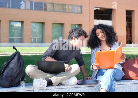 Deux étudiants latins étudiant à partir de leurs notes de cours assis sur un mur à l'extérieur. La vie universitaire sur le campus. Banque D'Images