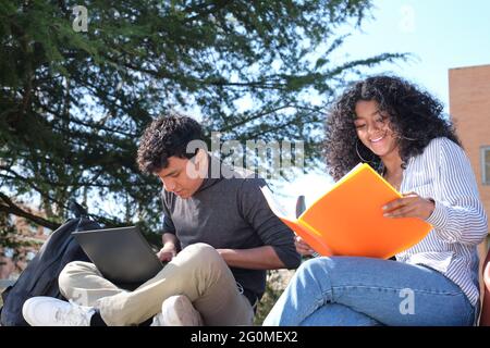 Deux étudiants latins étudiant à partir de leurs notes de cours assis sur un mur à l'extérieur. La vie universitaire sur le campus. Banque D'Images