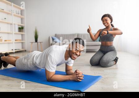 Sportif noir mec debout dans la posture de planche et souriant à l'appareil photo, sa petite amie le chroniant sur la montre intelligente, montrant le pouce vers le haut Banque D'Images