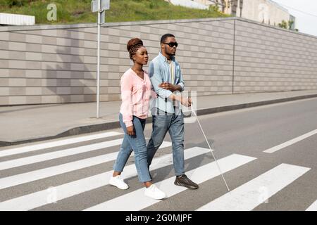 Jeune femme noire aidant un millennium malvoyants avec la canne traversant la rue de la ville. Concept de déficience visuelle Banque D'Images