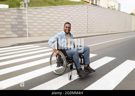 Pleine longueur d'homme noir handicapé dans la rue de passage en fauteuil roulant, en utilisant des passages en croix à l'extérieur Banque D'Images