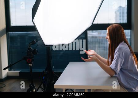 Vue latérale de la jeune femme blogger réussi enregistrement vidéo vlog en utilisant la caméra professionnelle et softbox assis au bureau sur l'arrière-plan de la fenêtre. Banque D'Images