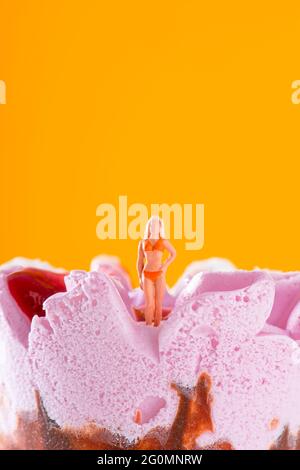 une femme miniature portant un bikini orange debout sur un cône de glace prérempli de fraise, sur un fond orange avec un peu d'espace sur t Banque D'Images