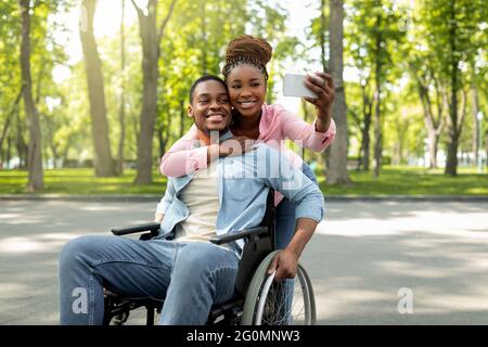 Une femme noire heureuse et son petit ami handicapé en fauteuil roulant emportant le selfie ensemble, se tenant dans le parc de la ville Banque D'Images