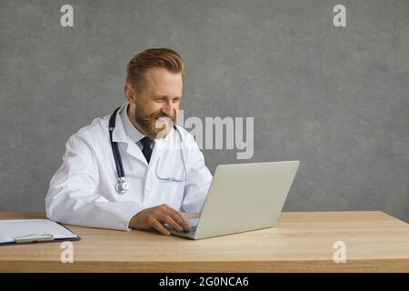 Joyeux médecin caucasien travaillant sur un ordinateur portable assis à la table sur un fond gris. Banque D'Images
