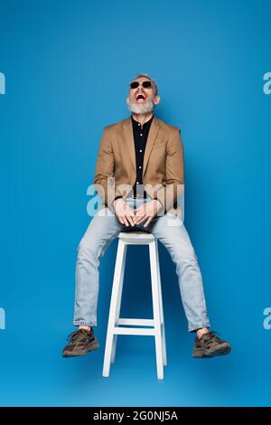 pleine longueur d'homme d'âge moyen en lunettes de soleil riant sur chaise blanche sur bleu Banque D'Images