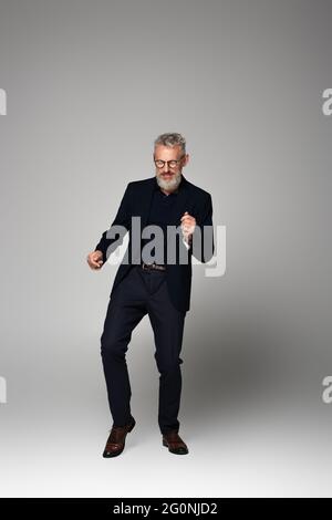 pleine longueur d'homme heureux d'âge moyen en lunettes et costume dansant sur gris Banque D'Images
