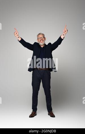 pleine longueur d'homme d'âge moyen souriant en lunettes et costume pointant avec les doigts sur le gris Banque D'Images