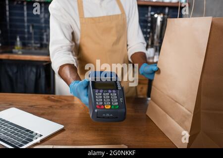 Vue rognée du barista afro-américain dans des gants en latex tenant le terminal de paiement et un sac en papier dans un café Banque D'Images