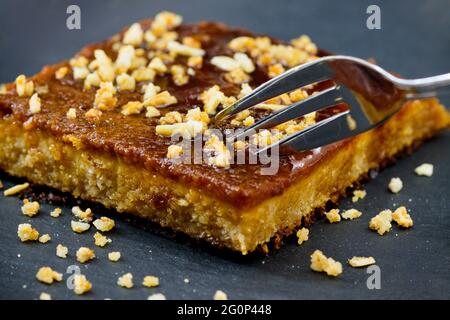 Quelqu'un coupe une portion d'un gâteau au biscuit avec du caramel et des morceaux d'amandes sur le dessus avec une fourchette sur une assiette noire Banque D'Images