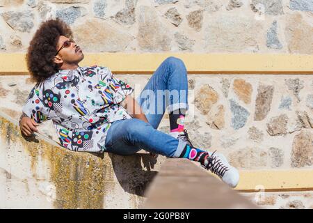 image horizontale d'un homme noir avec des cheveux afro et des lunettes rouges couchés sur le côté sur un mur en béton avec un mur en pierre en arrière-plan Banque D'Images